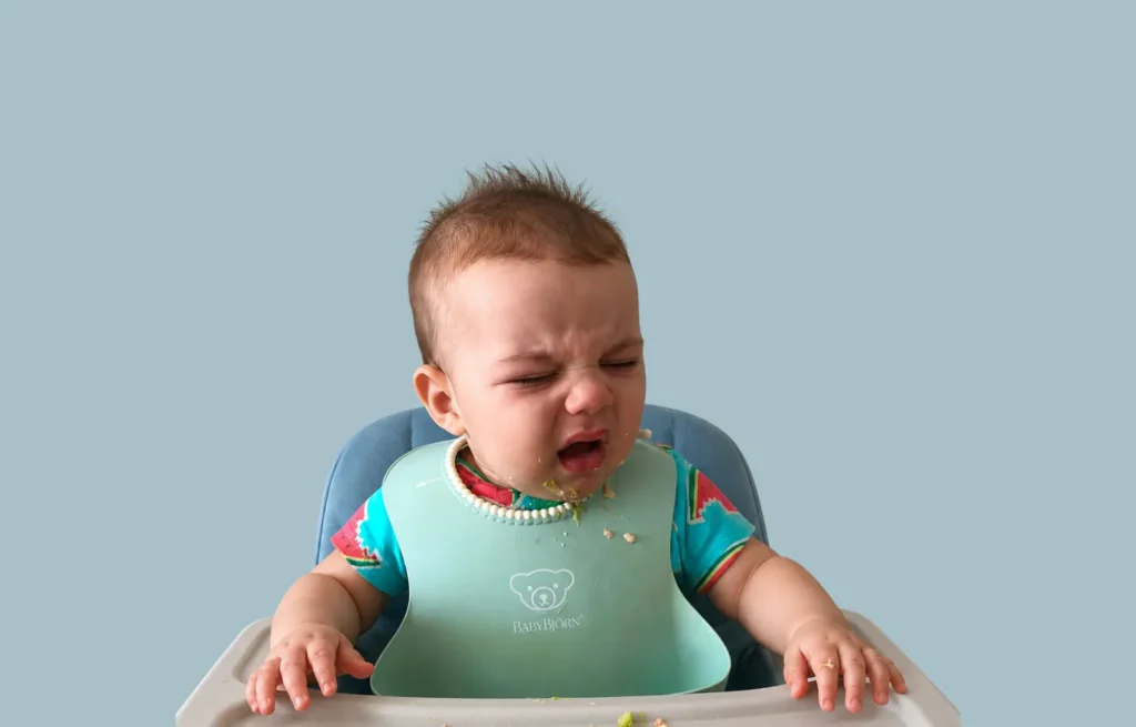 Reflexo de gag: bebê chora com os olhos fechados sentado na cadeira de alimentação, usando babador azul, com resquícios de comida no rosto e na bandeja