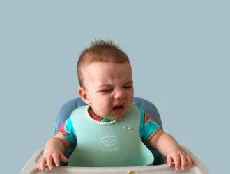Reflexo de gag: bebê chora com os olhos fechados sentado na cadeira de alimentação, usando babador azul, com resquícios de comida no rosto e na bandeja