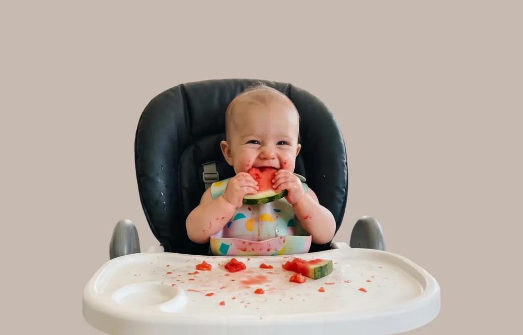 Bebê na cadeira de alimentação comendo uma fatia de melancia com as mãos durante a introdução alimentar do bebê