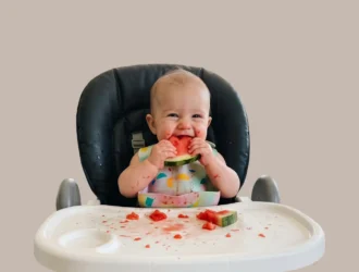 Bebê na cadeira de alimentação comendo uma fatia de melancia com as mãos durante a introdução alimentar do bebê